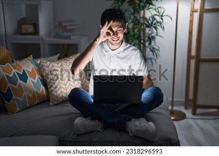 Young hispanic man using laptop at home at night doing ok gesture with hand smiling, eye looking through fingers with happy face. 
