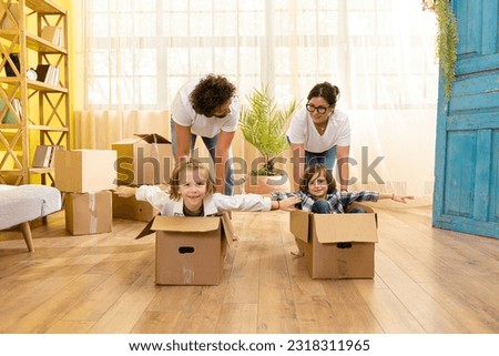 In front of the camera after young family moving to new home they take their kids and put them on the boxes and start to doing a ride through the living room Royalty-Free Stock Photo #2318311965