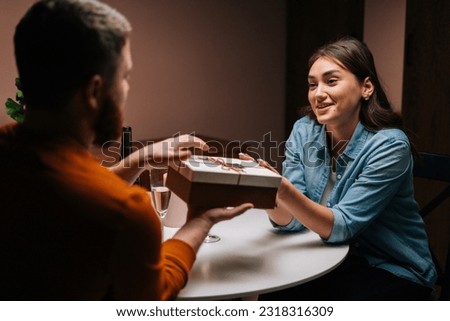 Attractive young woman giving wrapped box with gift to unrecognizable man at table with candles on birthday or Valentines Day. Happy husband receiving present from wife enjoying romantic dinner date.