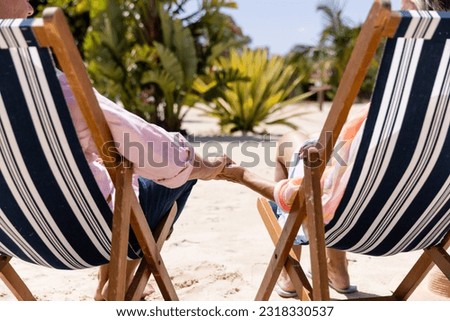 Low section of caucasian romantic senior couple holding hands and sitting on deck chairs at beach. Copy space, unaltered, retirement, vacation, love, together, enjoyment, relaxing nature and summer. Royalty-Free Stock Photo #2318330537