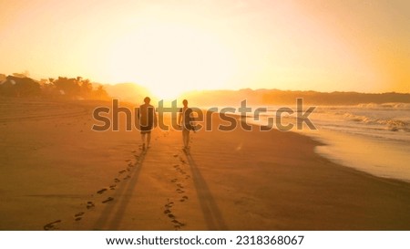 SILHOUETTE: Surf couple walking along gorgeous sandy beach towards golden sunset. Two friends on a holiday surf trip at Playa Venao. They are going surfing and riding waves in beautiful sunset light. Royalty-Free Stock Photo #2318368067