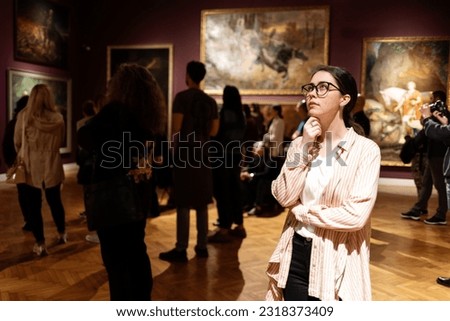 Thoughtful young Caucasian woman wearing glasses and looking at exhibition. In background, people are looking at paintings. Concept of Museum Day Royalty-Free Stock Photo #2318373409