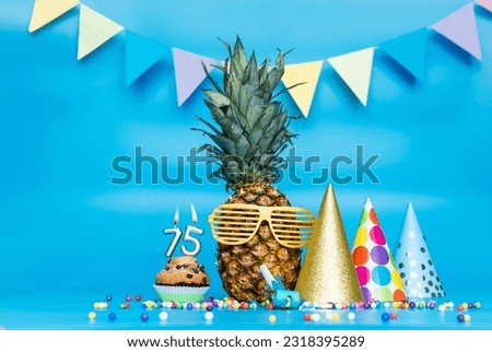 Creative background with pineapple character in sunglasses copy space. Happy birthday background with muffin or cake with candle number  75. Anniversary holiday decorations on a blue background.