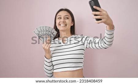 Young beautiful hispanic woman taking selfie holding dollars over isolated pink background