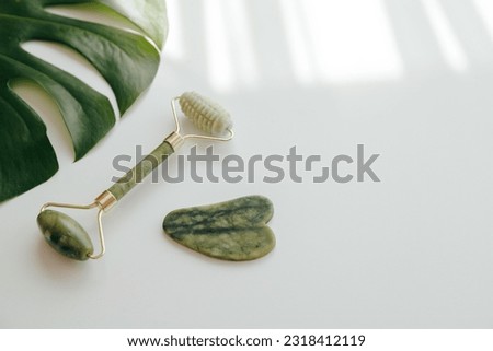 Green Facial Jade Massage Roller and Gua Sha Massager with Monstera Leaf on white background.