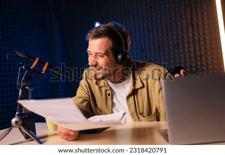 Smiling and gesturing radio host with headphones reading news from paper into studio microphone at radio station with neon lights