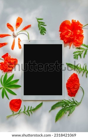 Digital tablet and poppy flowers composition on a light gray background with sunlight and shadows.
