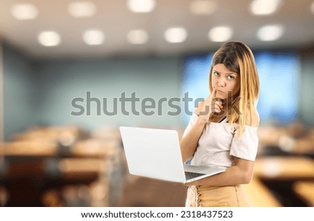 Curious blonde teenager girl thinking while holding a laptop computer against a classroom background