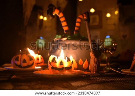 Pumpkin Jack candles. Funny witch cauldron cake with broom, green sweet bubbles, and two legs in orange striped stockings and heavy shoes. Orange lights in the room at Halloween home party for kids.