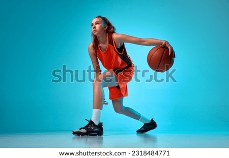 Young athletic girl, basketball player in motion, running with ball against blue studio background in neon light. Concept of professional sport, action and motion, game, competition, hobby, ad
