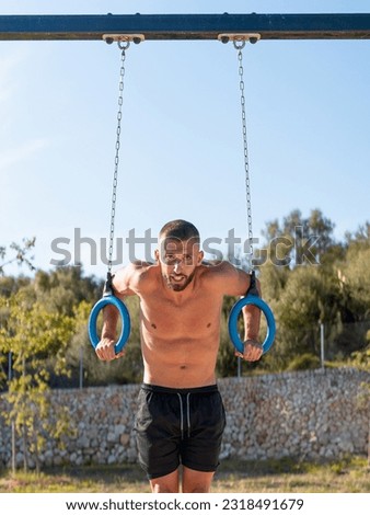 Vertical photo of a shirtless young man exercising with the rings at the outdoor calisthenics park - calisthenics concept