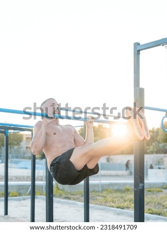 vertical photo of young shirtless athletic man performing the l-sit exercise on the bar of the outdoor calisthenics park at sunset - calisthenics concept
