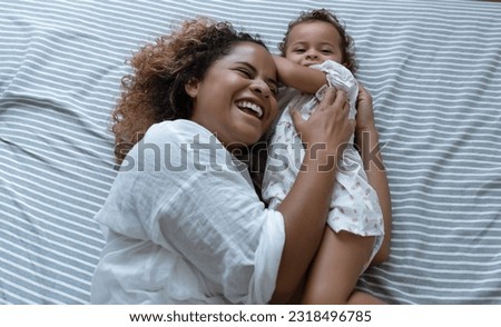 Happy African American Mother and adorable daughter relaxing and playing on the bed at the weekend together, lazy morning, warm and cozy scene. Having fun enjoy funny moments in bedroom.