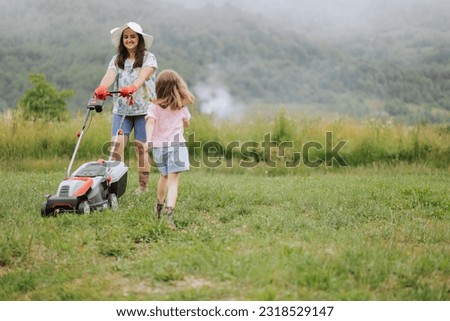 A woman in boots with her child in the form of a game mows the grass with a lawnmower in the garden against the background of mountains and fog, garden tools concept, work, nature.