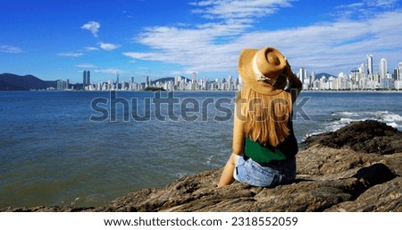 Panoramic banner view of young woman sitting on the rocks looking at Balneario Camboriu skyline on Atlantic Ocean. Summer vacation in Southern Brazil.
