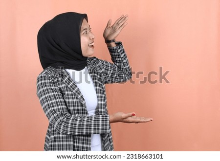Smiling beautiful Asian muslim woman pointing finger to empty space wearing a striped gray jacket and veil isolated peach background