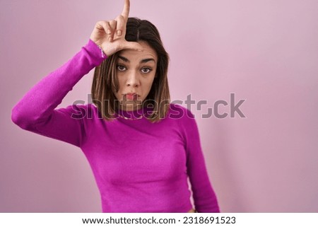 Hispanic woman standing over pink background making fun of people with fingers on forehead doing loser gesture mocking and insulting. 