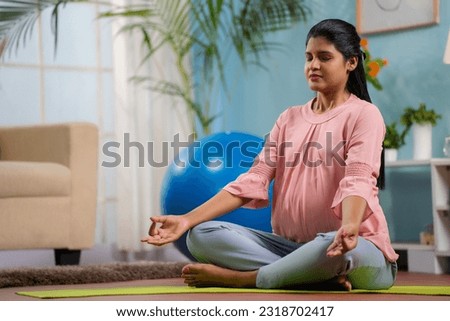 Indian peaceful pregnant woman in lotus pose doing meditation on yoga mat home - concept of relaxation, healthy pregnancy and self-care. Royalty-Free Stock Photo #2318702417