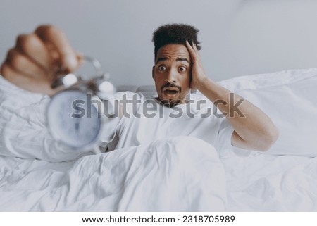 Young late awake disappointed sad african american man in nightwear hold in hand clock alarm lying in bed rest relax spend time in bedroom lounge home in own room house wake up. Morning rush concept Royalty-Free Stock Photo #2318705989