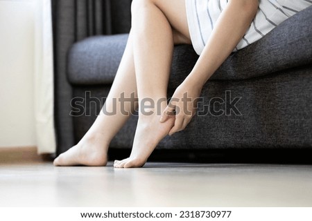 Legs of young woman,girl sitting on sofa holding foot with hand,feeling pain and stiffness in sole of foot and heel bone while standing,painful heel problem caused by Plantar Fasciitis and Flat Feet