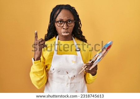 African woman holding painter palette pointing up looking sad and upset, indicating direction with fingers, unhappy and depressed. 