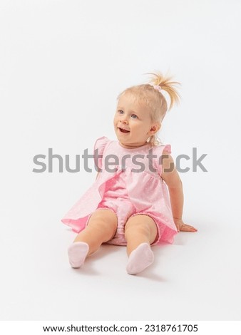 Surprise, delight.A cute blonde girl 1-2 years old in a pink dress is sitting on the floor with her mouth open on a white background. Copy space.