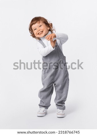 A cheerful 2-year-old toddler with curly hair is indulging, laughing, smiling, holding his hands to himself and bending over in a gray jumpsuit and a white turtleneck Royalty-Free Stock Photo #2318766745