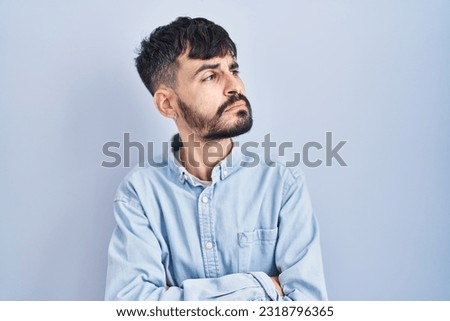 Young hispanic man with beard standing over blue background looking to the side with arms crossed convinced and confident 