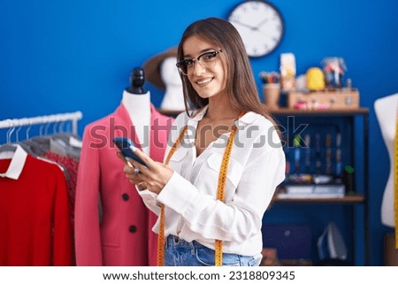 Young beautiful hispanic woman tailor smiling confident using smartphone at clothing factory