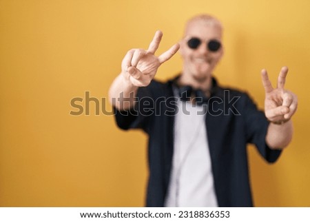 Young caucasian man wearing sunglasses standing over yellow background smiling with tongue out showing fingers of both hands doing victory sign. number two. 