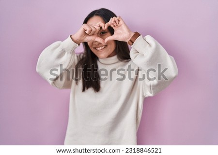 Young south asian woman standing over pink background doing heart shape with hand and fingers smiling looking through sign 