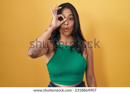 Brunette woman standing over yellow background doing ok gesture shocked with surprised face, eye looking through fingers. unbelieving expression. 