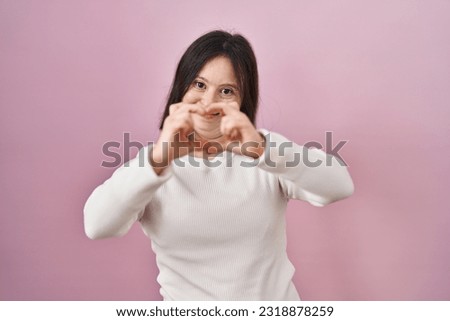 Woman with down syndrome standing over pink background smiling in love doing heart symbol shape with hands. romantic concept. 
