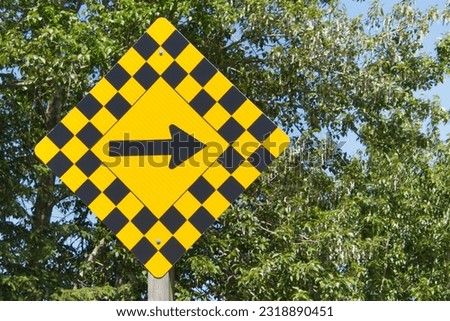 Yellow and black road sign pointing to the right, showing turn ahead. Green foliage in background.