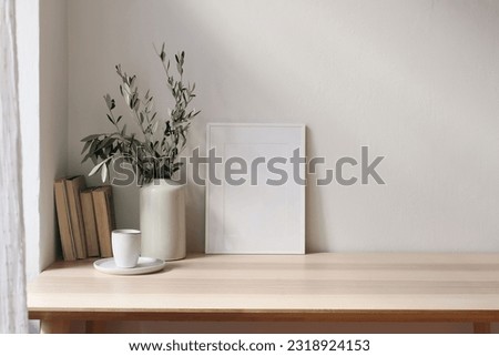 Summer home, elegant interior. Ceramic vase with olive tree branches, cup of coffee. Blank white picture frame mockup on wooden table. Window with white curtains. Living room. Mediterranean design.