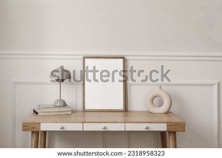 Elegant living room, home office. Modern round vase, beige lamp and old books. Blank wooden picture frame mockup. Bamboo table with white drawers. Scandi boho interior, home. Beige wall background.