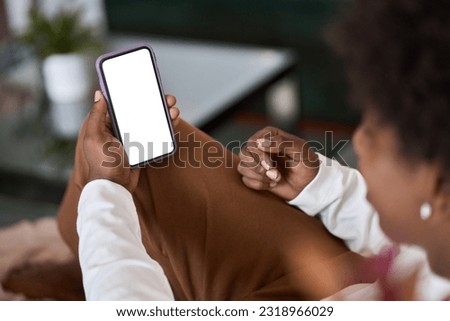 Over shoulder view of gen z African American teen holding smartphone mock up white cell screen template advertising mobile application, watching social media videos on cellphone at home on couch.