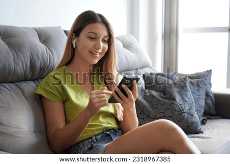 Happy young woman hanging on social media with phone sitting on sofa at home