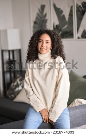 Young adult smiling pretty hispanic curly woman posing at home looking at camera, happy calm beautiful positive woman in modern cozy apartment living room, close up headshot vertical portrait.