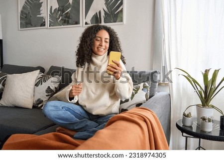 Smiling latin girl sitting on couch using cell phone in living room at home. Relaxed happy young woman holding mobile cellphone technology doing shopping, communicating online, watching social media. Royalty-Free Stock Photo #2318974305
