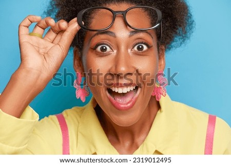 Close up shot of young black very pleased girl smiling broadly and taking off her glasses posing against blue wall, happy time concept, copy space