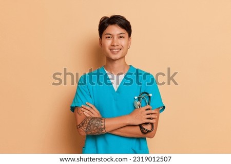Young smiling male medical worker holds stethoscope in hand standing with his arms crossed, poses on studio background, health care concept, copy space