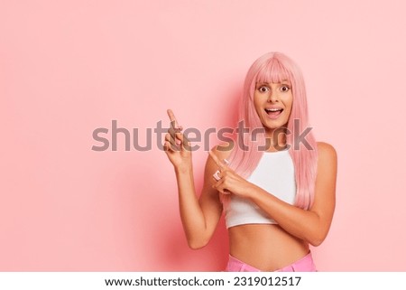 Young pretty woman stands against pink background, pointing index fingers aside with smile, wearing white top, pleasure concept, copy space Royalty-Free Stock Photo #2319012517