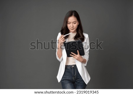 Asian female executive with long hair lead the organization holding and looking at tablet Raise the pen and think carefully. She wore a white suit, jeans and stood in a gray studio setting. Royalty-Free Stock Photo #2319019345