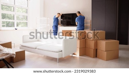Two Young Male Movers In Uniform Placing Television In Living Room Royalty-Free Stock Photo #2319029383