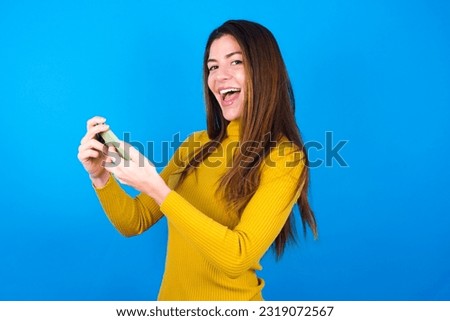 Nice addicted cheerful young beautiful woman wearing yellow sweater over blue studio background using gadget playing network game