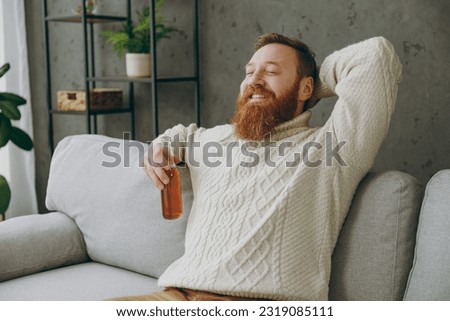 Young ginger man wear casual clothes drink ale hold hands behind neck close eyes sits on grey sofa couch stay at home hotel flat rest relax spend free spare time in living room indoor. Lounge concept Royalty-Free Stock Photo #2319085111