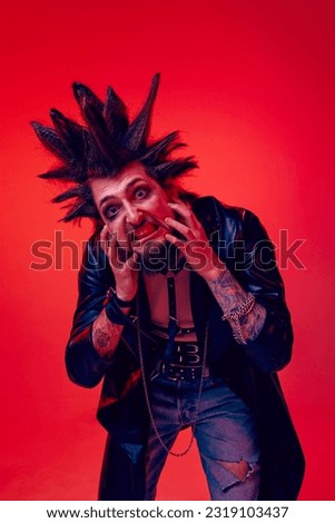 Crazy face. Portrait of expressive young man, punk posing in makeup and leather jacket against red studio background in neon light. Concept of music, lifestyle, subculture, art, youth, human emotions