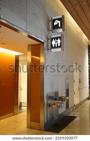 Sign direction to disabled, lady and gentleman toilet, drinking water in airport terminal building. View of male female bathroom doors across from each other drinking water fountain mounted on wall.