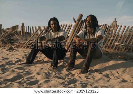 Portrait of two African men wearing dreadlocks on ocean posing during sunset sitting near fence on dunes